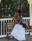Woman sitting on a bench holding a drink, with trees and a white railing in the background